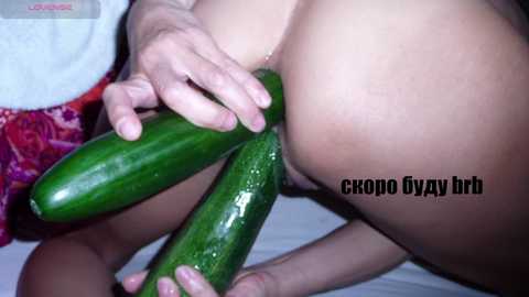 Video of a light-skinned woman with a plump, shaved vulva, holding four large green cucumbers, creating an explicit, erotic scene.