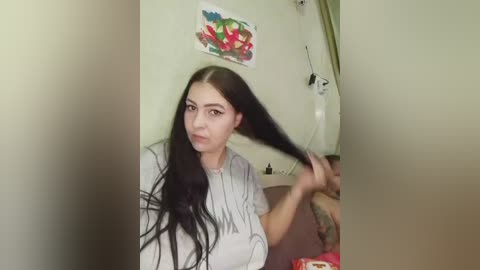 Video of a young woman with long, straight black hair, holding her hair while wearing a light grey shirt, standing in a dimly lit room with green walls and a colorful painting on the wall.