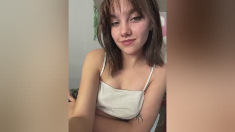 Media: Video of a young woman with fair skin, straight brown hair, and a white crop top, sitting indoors with a blurred background.
