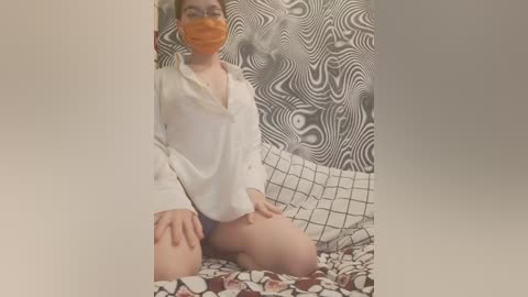 A video of a fair-skinned, short-haired woman with glasses and an orange face mask, kneeling on a bed with black-and-white floral bedding. She wears a white blouse and has her hands on her knees.
