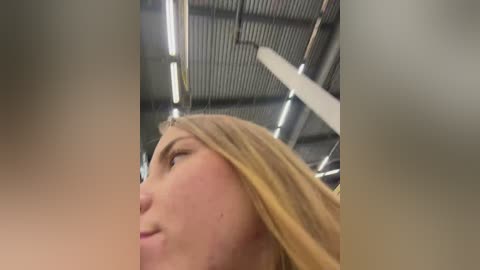 Media: Video of a close-up side profile of a young woman with long blonde hair, fair skin, and light makeup, captured indoors with metallic ceiling panels and fluorescent lights in the background.