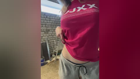 Video of a woman in a red T-shirt and grey sweatpants, standing in a cluttered room with a brick wall, a white chair, and a blue and white dog bowl.