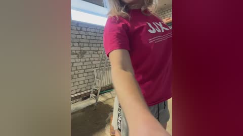 Video of a woman with light skin, blonde hair, wearing a red T-shirt with white text, standing in a room with a brick wall and exposed concrete floor.