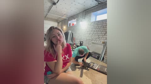 A video of a young woman with long blonde hair, wearing a red t-shirt, sitting on a wooden floor in a cluttered workshop with exposed brick walls and fluorescent lighting.