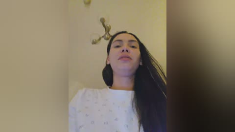 Video of a young woman with long black hair and fair skin, wearing a white hospital gown, standing against a beige wall with a golden light fixture.