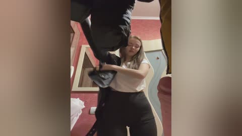 Media: Video of a woman in a white top and black pants struggling to remove a man's black jacket, while another person's leg is visible in the foreground. The background shows red walls, a mirror, and a white towel.