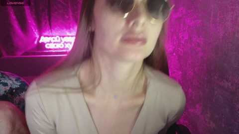 Video of a fair-skinned woman with long, straight brown hair, wearing oversized sunglasses, a light-colored V-neck top, and a floral-patterned pillow behind her, with a pink and purple neon-lit background.