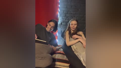 Media: Video of two people: a man with glasses, reading a book, and a woman with long hair, smiling, sitting on a patterned cushion against a brick wall with red and blue lights.