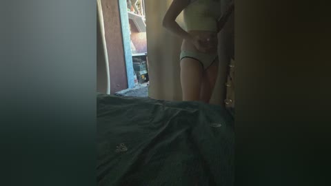 A video of a slender, light-skinned woman with a flat stomach and small breasts, wearing a green crop top and high-waisted panties, standing in a dimly lit room. The background reveals a cluttered space with a bed covered in dark sheets.
