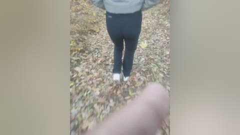 Media: A video captures a person's back in tight black pants and a gray jacket, standing on a ground covered in fallen leaves, with a blurred hand in the foreground.