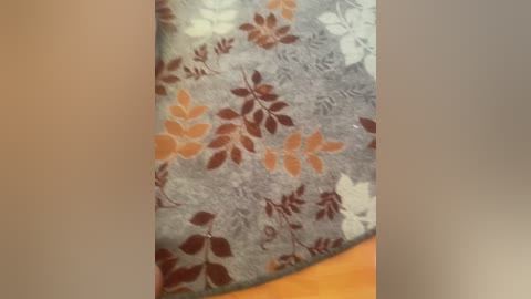 Video of a gray fabric with a floral pattern featuring orange, brown, and white leaves, partially covering a wooden surface. The fabric appears smooth and soft, possibly a throw or blanket.