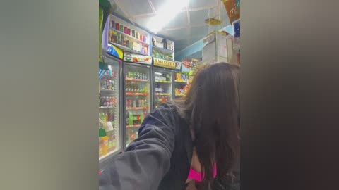 Media: Video of a crowded convenience store with a woman in the foreground, partially obscured, selecting items from shelves. Brightly lit, colorful displays of snacks and beverages fill the background.