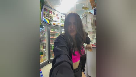 Media: Video of a woman with long dark hair and a pink bra, standing in a brightly lit convenience store. Shelves are stocked with beverages and snacks, and a ceiling fan is visible.