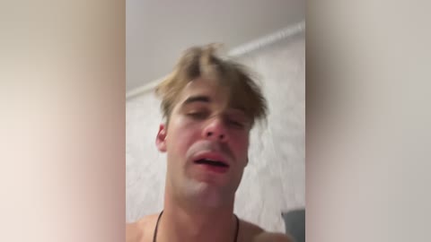 Video of a shirtless young man with tousled light brown hair, closed eyes, and a slightly open mouth, standing in front of a white textured wall.