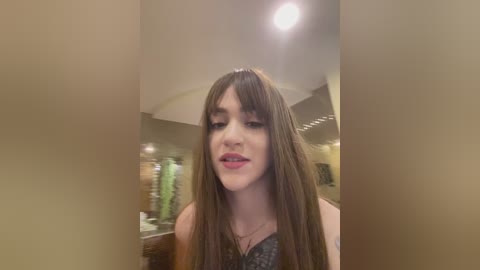 Media: Video of a young woman with long, straight brown hair, wearing a dark patterned top, smiling, in a modern bathroom with beige walls and a light fixture.
