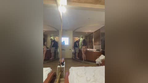 Video of a modern hotel room with mirrored walls, featuring a man in a dark shirt and shorts adjusting the bed. The room has a wooden floor, white linens, and a TV on the wall.