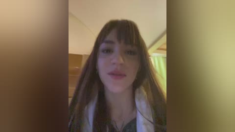 Media: A blurry video of a young woman with long, dark hair, fair skin, and a neutral expression, wearing a white shirt, standing in a dimly lit room with a green curtain in the background.