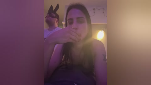 A video of a young woman with long brown hair, wearing a black bunny mask and a sleeveless top, blowing a kiss to a man in a white t-shirt. The room is dimly lit with purple and yellow hues, creating a playful, intimate atmosphere.