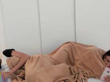 Video of a woman lying on her side, partially covered by a tan blanket, holding a pink phone in her right hand, with a white wall in the background.