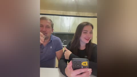 Media: Video of a man in a blue polo shirt and a woman in a black top, both smiling, taking a selfie in a modern kitchen with white cabinets and a stainless steel sink.