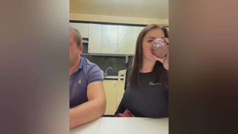 Media: Video of a middle-aged man in a blue polo shirt and a young woman with long brown hair in a black shirt, drinking from a glass in a modern kitchen with white cabinets.