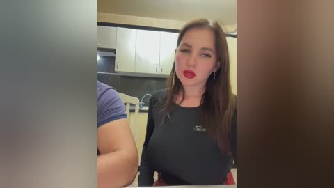 Media: Video of a Caucasian woman with long brown hair, red lipstick, and a black shirt, sitting in a modern kitchen with white cabinets and stainless steel appliances.