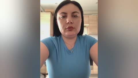 Video of a middle-aged woman with medium skin tone, straight black hair, wearing a light blue short-sleeved top. She stands in a modern kitchen with light wood cabinets, holding a camera.