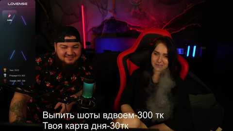 Media: Video of a bearded man in a floral shirt and cap, and a woman in a black fur coat, seated in red gaming chairs, in a dimly lit room with neon lights and glowing computer screens. Russian text overlay.