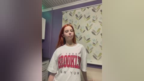 Video of a young woman with long, straight red hair wearing a white Brooklyn New York T-shirt, standing in a room with lavender walls and floral-patterned curtains.
