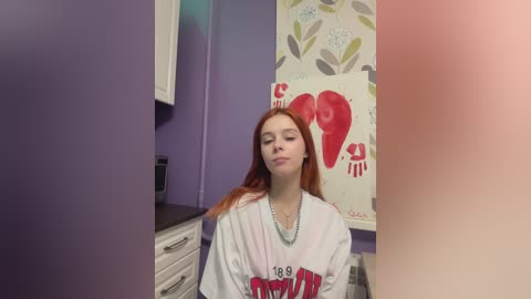 Video of a young woman with long red hair, wearing a white t-shirt with red text, sitting in a bathroom with purple and white walls.