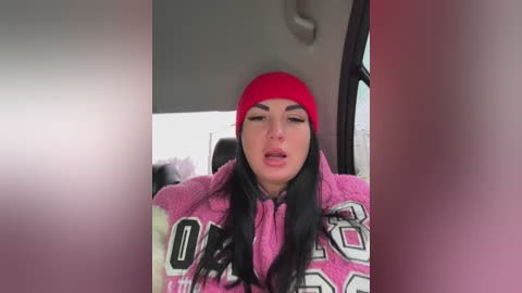 Video of a young woman with long black hair and fair skin, wearing a red beanie, pink hoodie with white letters, and sitting in a car. She appears relaxed, eyes closed, lips slightly parted.