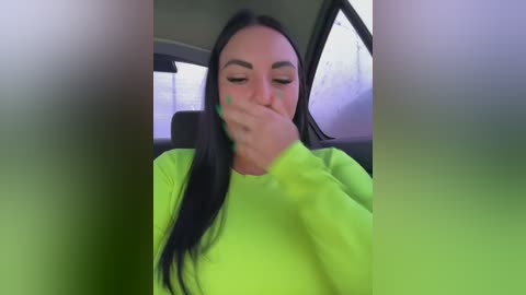 A video of a young woman with long black hair, wearing a bright yellow sweater, sitting in a car. She covers her mouth with her hand, looking tired and distressed. The car's interior is dimly lit.