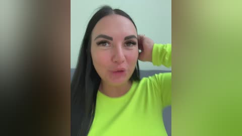 Video of a young woman with long black hair, fair skin, and large dark eyes, wearing a neon yellow long-sleeve top. She is sitting with one hand on her face, indoors with a blurred background.