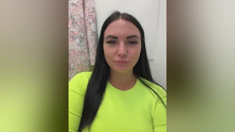 Video of a young woman with long black hair, fair skin, and wearing a bright green long-sleeved top. She stands in a bathroom with floral curtains and a white door in the background.