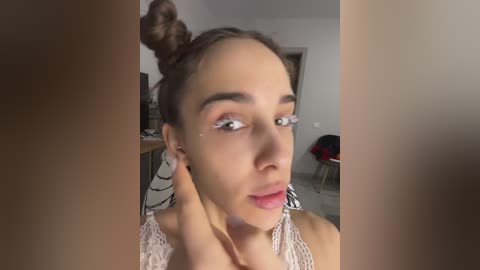 Media: Video of a young woman with light skin and brown hair in a topknot, wearing a white lace top. She has a neutral expression, with makeup including eyeshadow and pink lipstick. Background shows a simple room with a table, chair, and a black and white patterned chair.