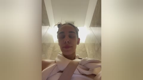 Media: Video of a woman with light brown skin, braided hair, and a white towel wrapped around her neck, taken from a low angle. The background features a beige tiled wall and a white ceiling.