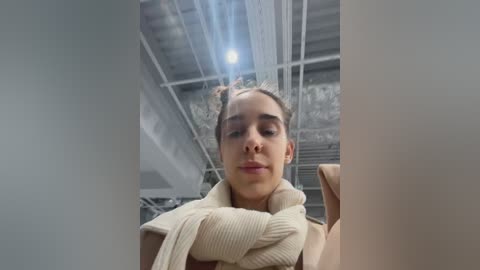 Media: Video of a young woman with fair skin and brown hair in a bun, wearing a beige sweater, standing indoors under a high ceiling with exposed beams and industrial lights.