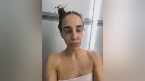 Media: A video of a young woman with light skin and dark brown hair in a messy bun, standing in a white-tiled bathroom, partially wrapped in a white towel, looking pensive.