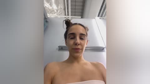 Media: Video of a young, light-skinned woman with dark hair in a messy bun, wearing a white towel, standing in a modern, industrial-style kitchen with exposed ductwork and a stainless steel fridge.