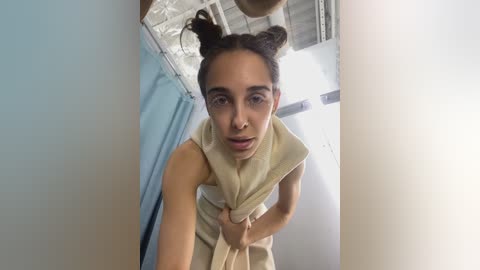 Media: Video of a young woman with light brown skin, dark brown hair in two buns, wearing a beige top, standing in a room with blue walls and white ceiling.