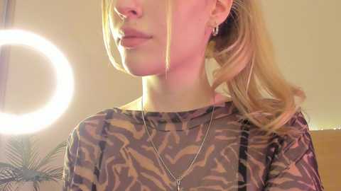 Media: Video of a blonde woman with fair skin, wearing a camouflage top, gold hoop earrings, and a silver necklace, captured in a softly lit room with a ring light.