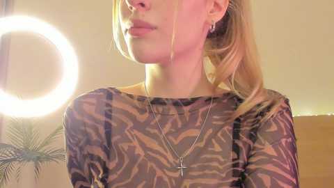 Video of a fair-skinned woman with blonde hair in a high ponytail, wearing a sheer, patterned top, a cross necklace, and large hoop earrings. Background shows a bright, circular light and a palm tree pattern.