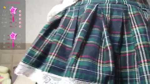 Media: Video of a person's lower back wearing a green plaid skirt with white lace trim, standing against a beige wall. The background includes a smartphone screen displaying a pink star emoji and text.