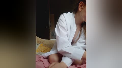Video of a woman in a white robe, sitting on a bed with yellow and pink bedding, partially obscured by a dark vertical gradient.