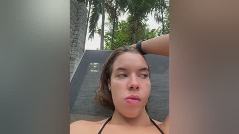 Media: Video of a young Caucasian woman with light brown hair, wearing a black bikini, posing outdoors in a tropical setting with palm trees and a stone wall.