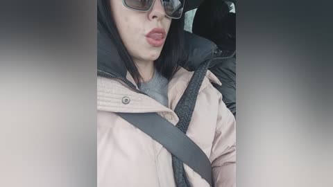 Media: Video of a woman with straight black hair, wearing black-framed glasses, a grey sweater, and a pink puffer jacket, with a seatbelt across her chest, seated in a car.