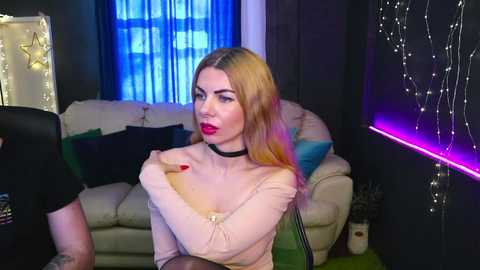 Video of a blonde woman with red lipstick, wearing a beige off-shoulder top, sitting in a dimly lit living room with blue curtains, a cream sofa, and a TV screen.