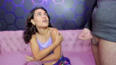 Video of a young woman with dark hair, wearing a purple tank top, kneeling on a pink tufted couch, looking up at a man's exposed, circumcised penis. Background features a honeycomb-patterned wall and blue lighting.