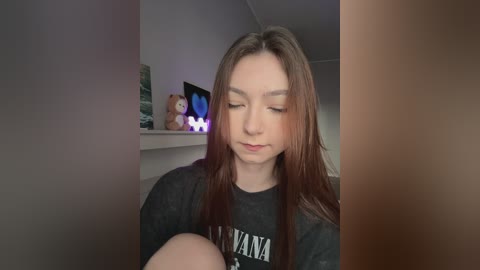 Media: A video of a young woman with long, straight brown hair, wearing a black Nirvana t-shirt, seated in a dimly lit room. She has a serene expression, eyes closed. Background includes a stuffed bear, a small glowing light, and a framed picture.