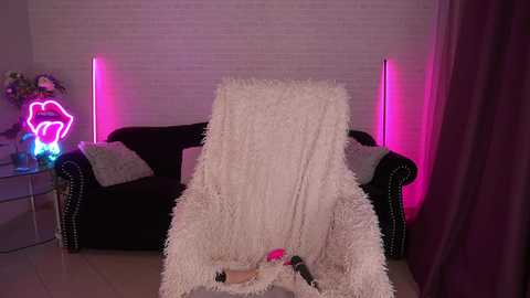 A video of a cozy living room with a plush white blanket draped over a dark velvet couch, illuminated by pink neon lights. A colorful neon heart-shaped sign is visible in the background.